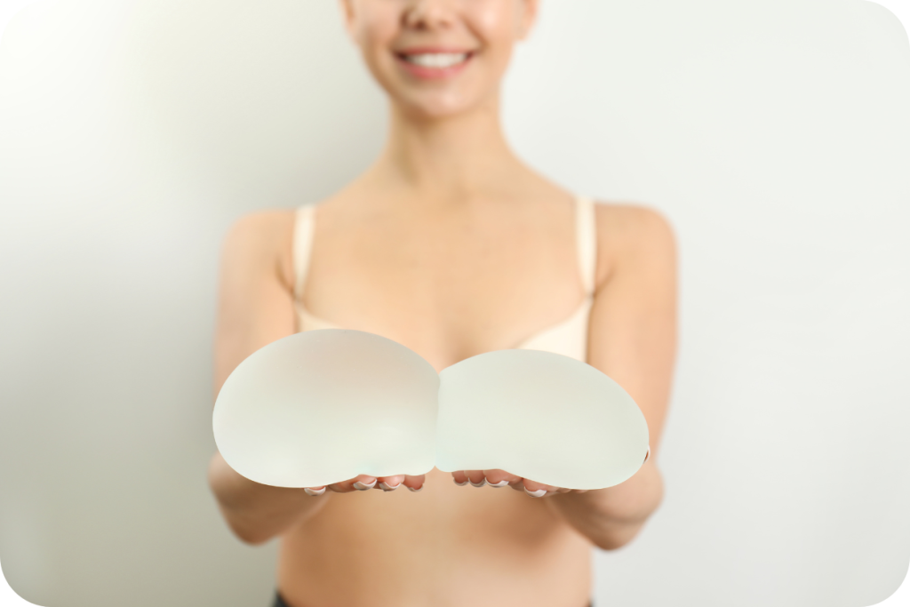 Imagem de uma mulher sorrindo, com o corpo desfocado do pescoço para baixo, segurando duas próteses de silicone opacas (implantes mamários) em primeiro plano, com as mãos à frente do peito, na altura do sutiã.