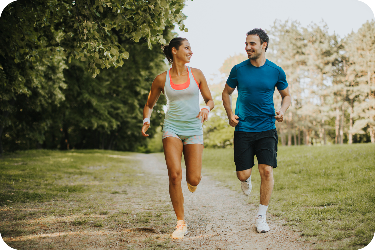 Duas pessoas, um homem e uma mulher, com aparência atlética e roupas esportivas, correndo e sorrindo em uma trilha ao ar livre, cercados por árvores e luz natural.