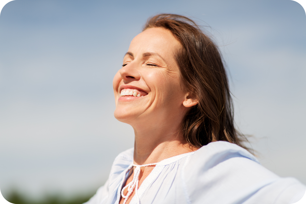 Close-up de uma mulher de meia-idade sorrindo com os olhos fechados sob o sol e céu azul, transmitindo sensação de liberdade, felicidade e renovação de autoestima, associada aos resultados do Mommy Makeover.
