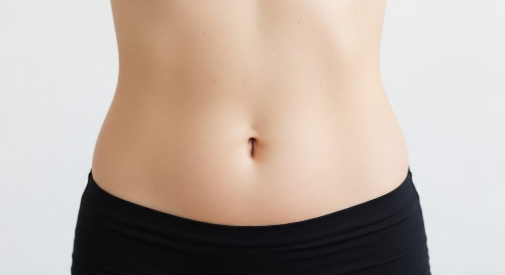 Close-up frontal do abdômen de uma pessoa com pele clara e saudável. A imagem destaca a região do umbigo e a linha da cintura, com a pessoa vestindo a parte superior de uma calça ou bermuda preta. O fundo branco e a iluminação suave enfatizam a textura natural da pele, ideal para ilustrar temas de cuidados corporais e estética.